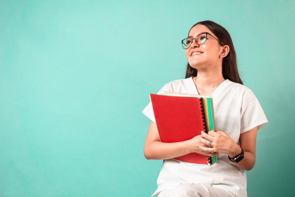 Portrait of a young, cheerful woman with glasses wearing white scrubs and holding folders